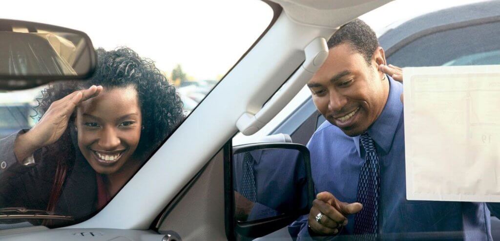 A black woman and black man look in car window with happy expressions on their faces
