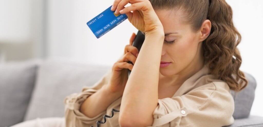 A white woman sits on a couch on the phone holding a credit card with a face of disappointment