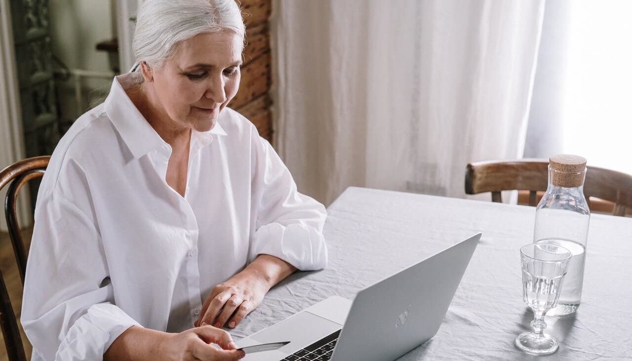 An older woman with white hair sits at a kitchen table looking at a laptop computer