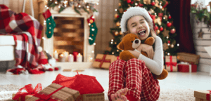 A little girl is very happy holding a teddy bear with a santa hat on her head. A christmas tree and fireplace are in the background