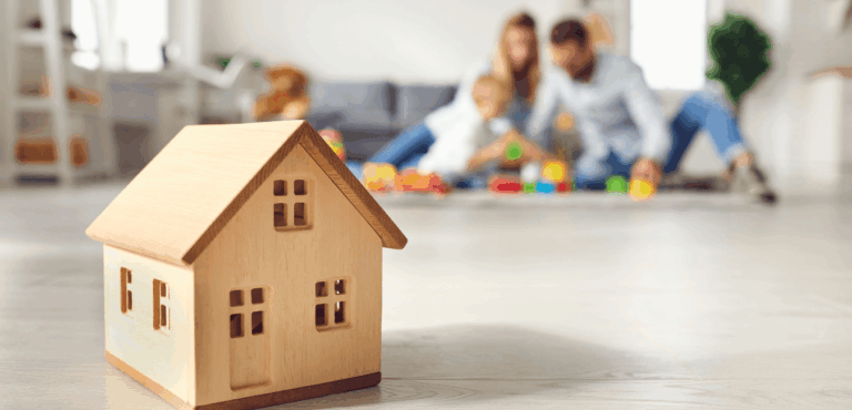 Close-up of a small wooden house model on the floor, with a joyful family playing together in the blurred background.