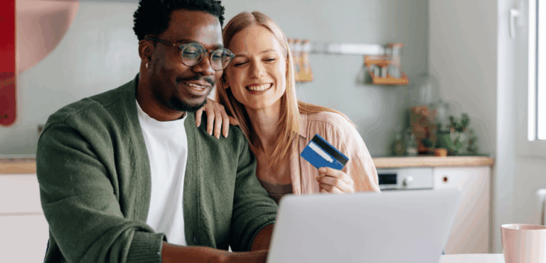 A couple sits at the kitchen table, smiling as they look at laptop screens while the woman holds a credit card.