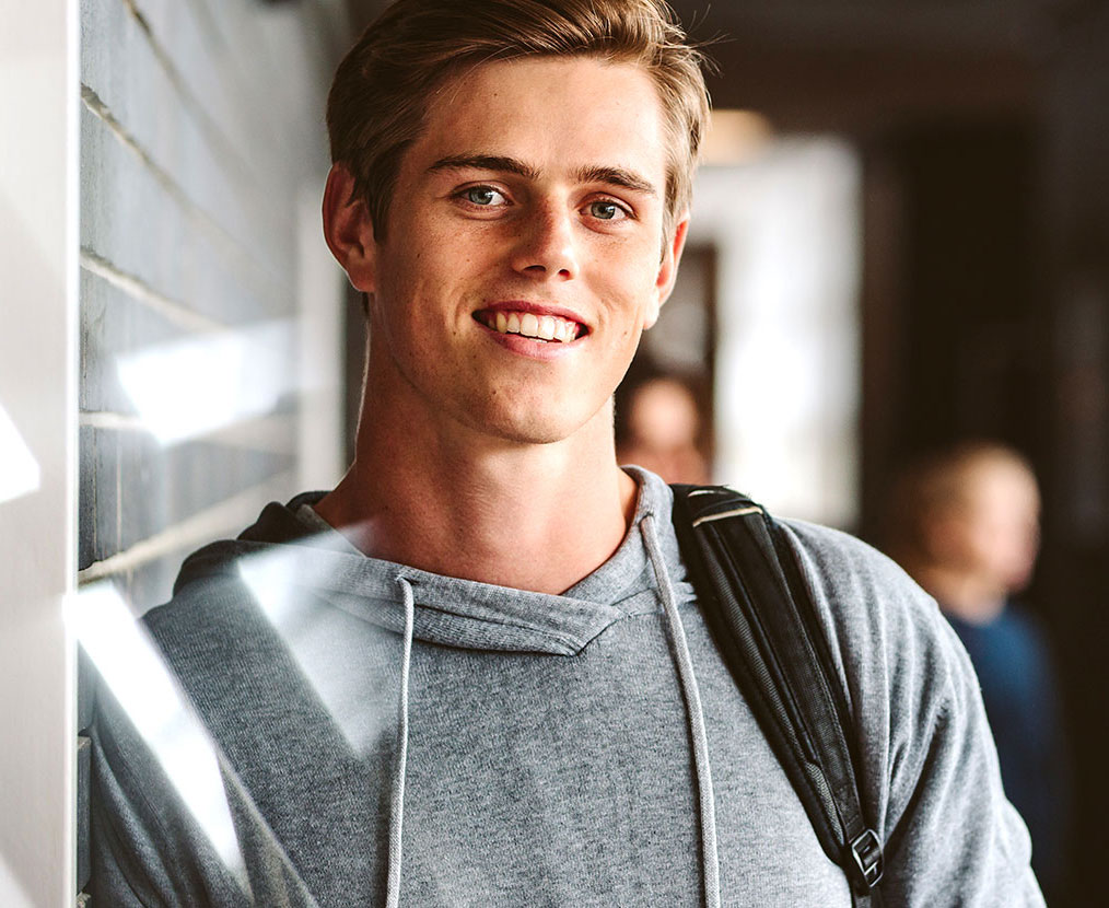 Smiling student wearing a gray hoodie and backpack in a school hallway
