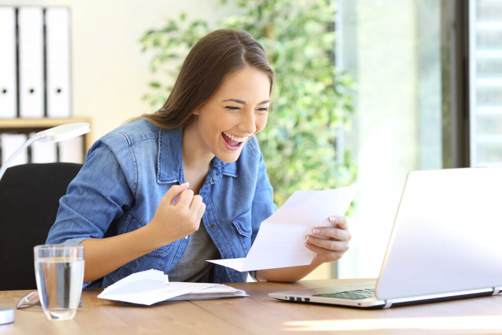 Smiling woman reading a letter at a table beside a laptop