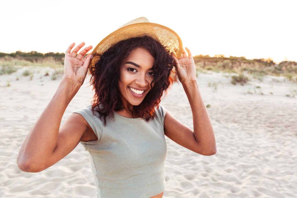 Smiling woman holding the brim of her straw hat on a sandy beach